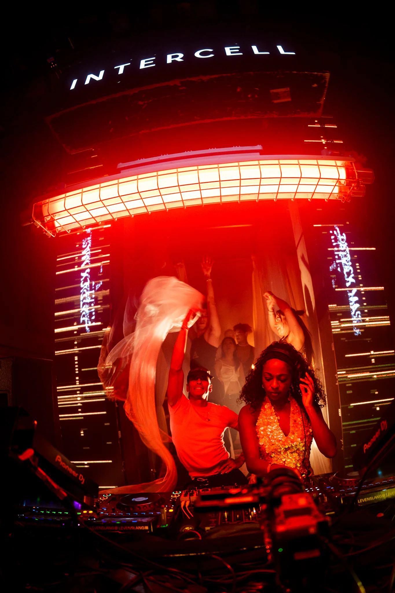 Two DJs performing beneath the Intercell sign in a red-lit booth during ADE