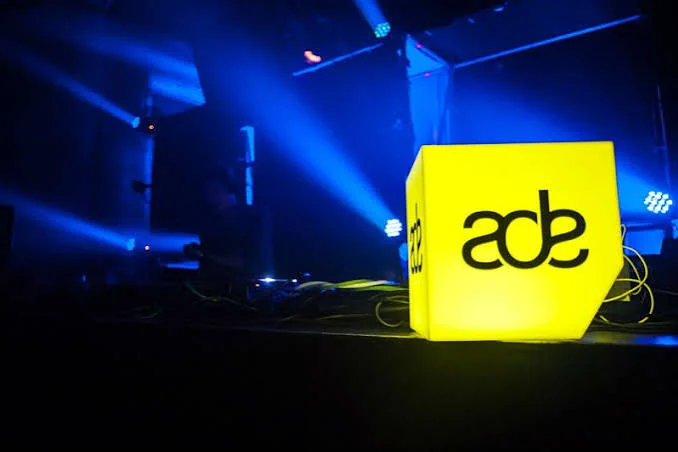 A glowing yellow ADE logo cube beside a dark DJ booth under blue lights
