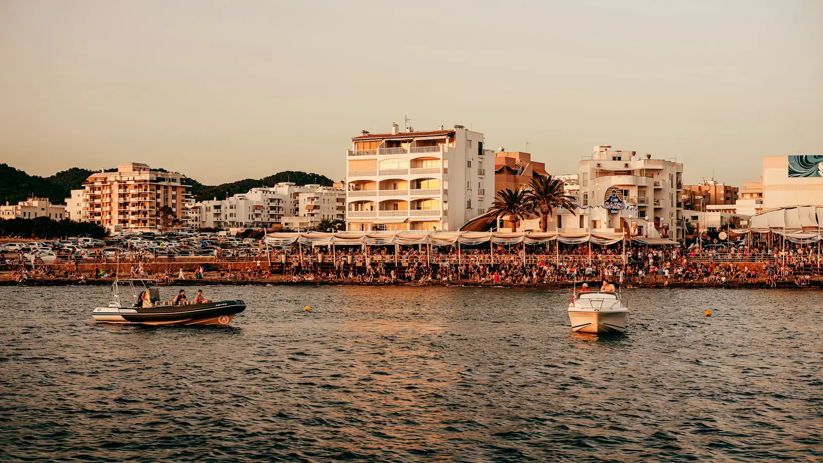 The sunset strip at San Antonio, Ibiza: a packed waterfront terrace under the Café Mambo sign at golden hour, with boats moored on the water in front of it