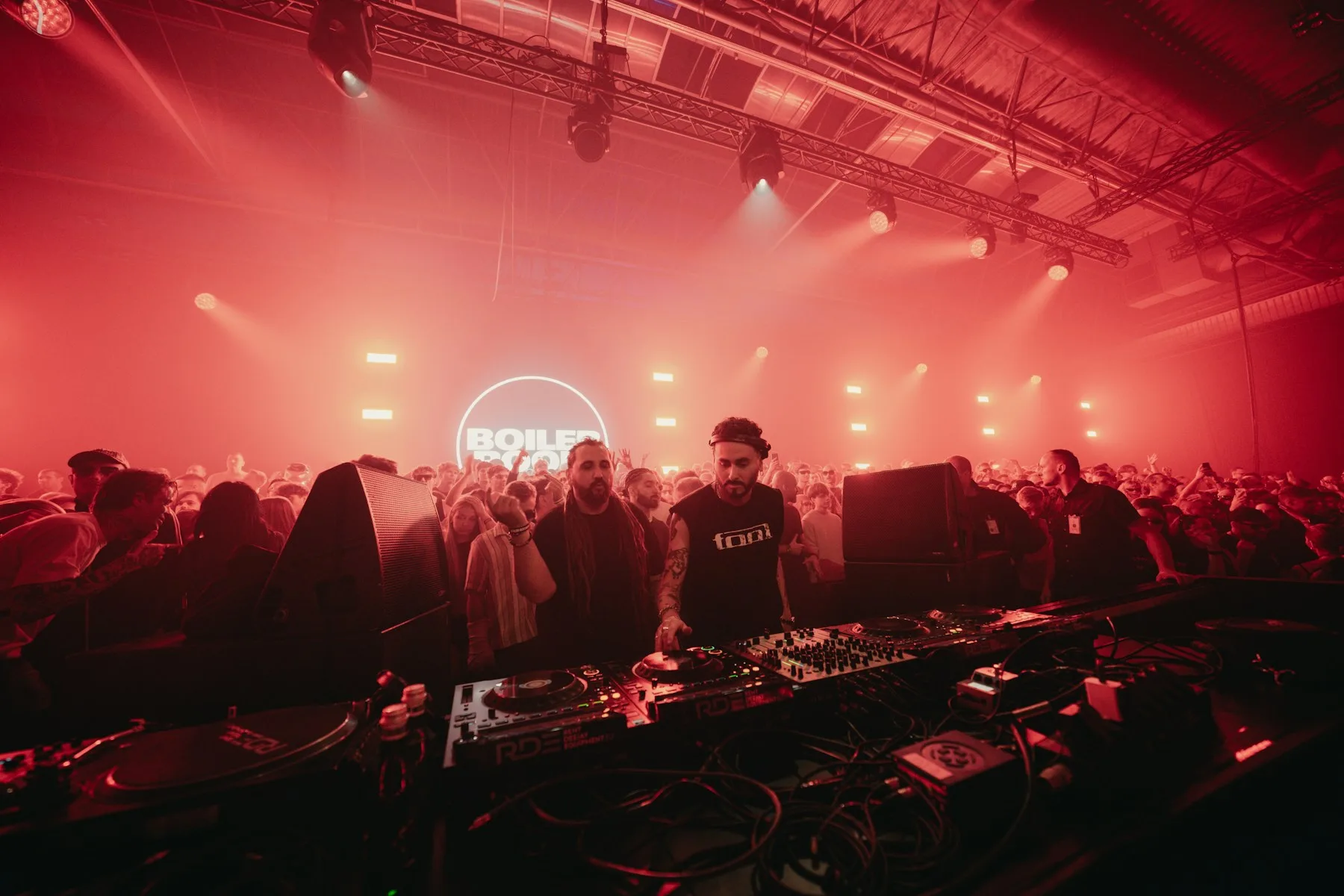 DJs at a mixing desk surrounded by a crowd under red lights, with a Boiler Room sign behind them