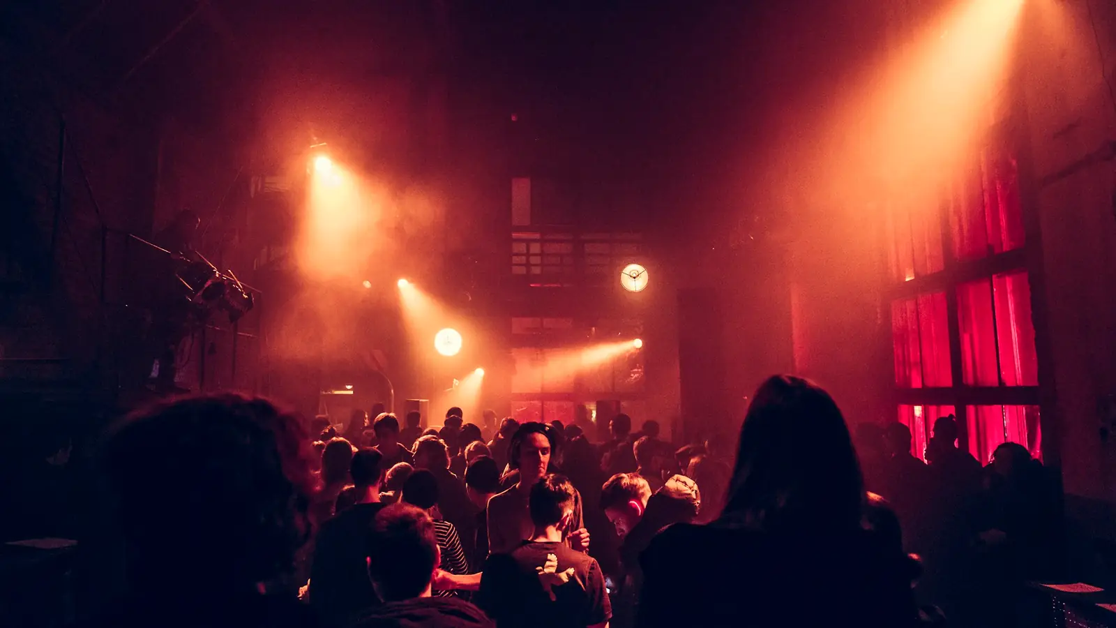 A hazy warehouse floor lit in warm orange, a crowd moving under a beam of light with red-curtained windows behind them
