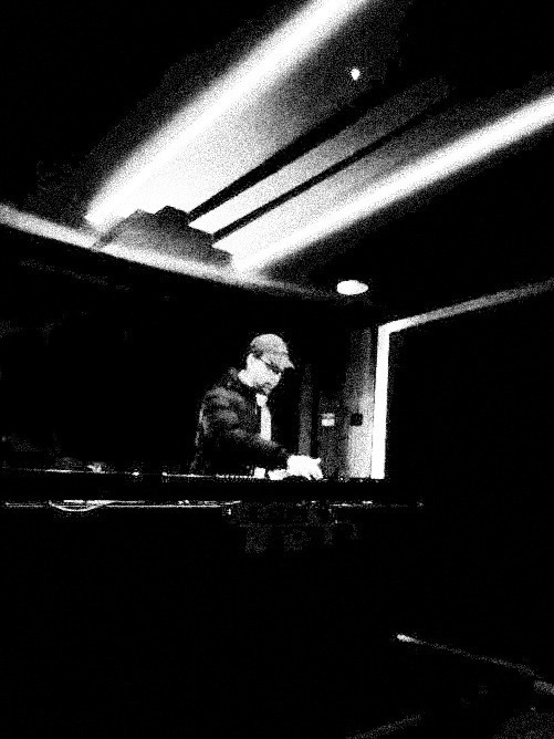 A dark studio room lit by ceiling strip lights, one person alone at the decks, no audience anywhere in the frame