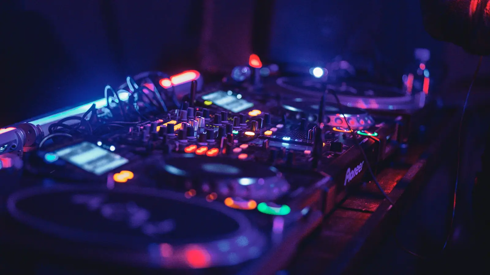 A booth in the dark: two CDJs either side of a mixer, lit by their own displays and one strip of blue light, headphones coiled on the deck