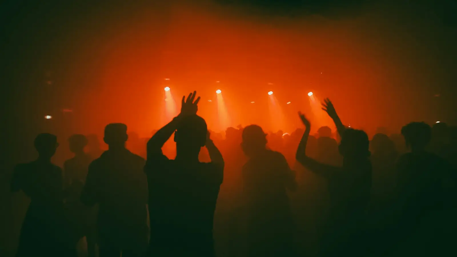 A dance floor in silhouette under orange haze, a few hands raised, the lamps at the far end of the room the only thing actually lit