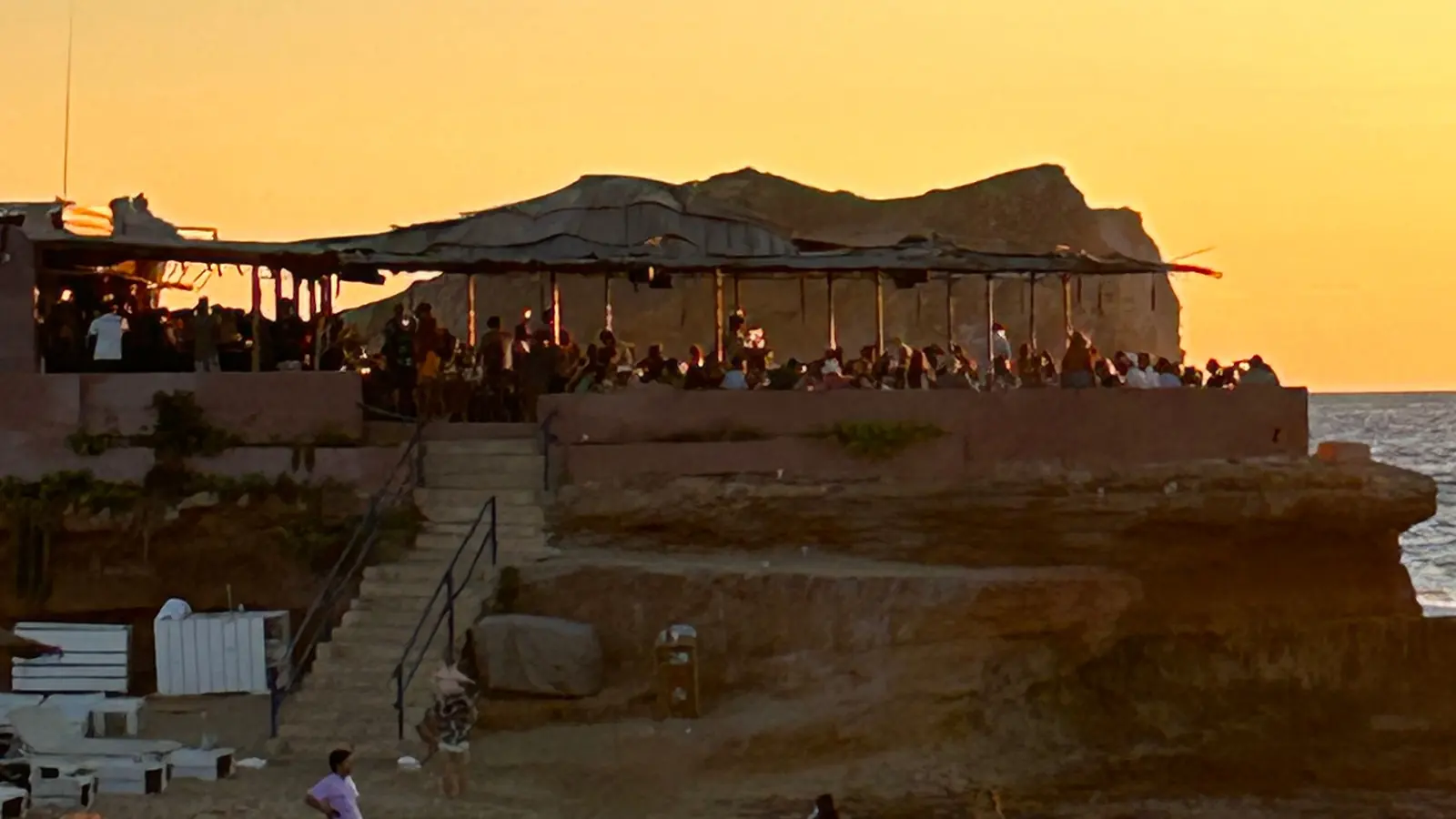 A packed open-air terrace under a slatted timber awning on a rocky headland at sunset, a cliff silhouetted behind it and steps down to a small beach below