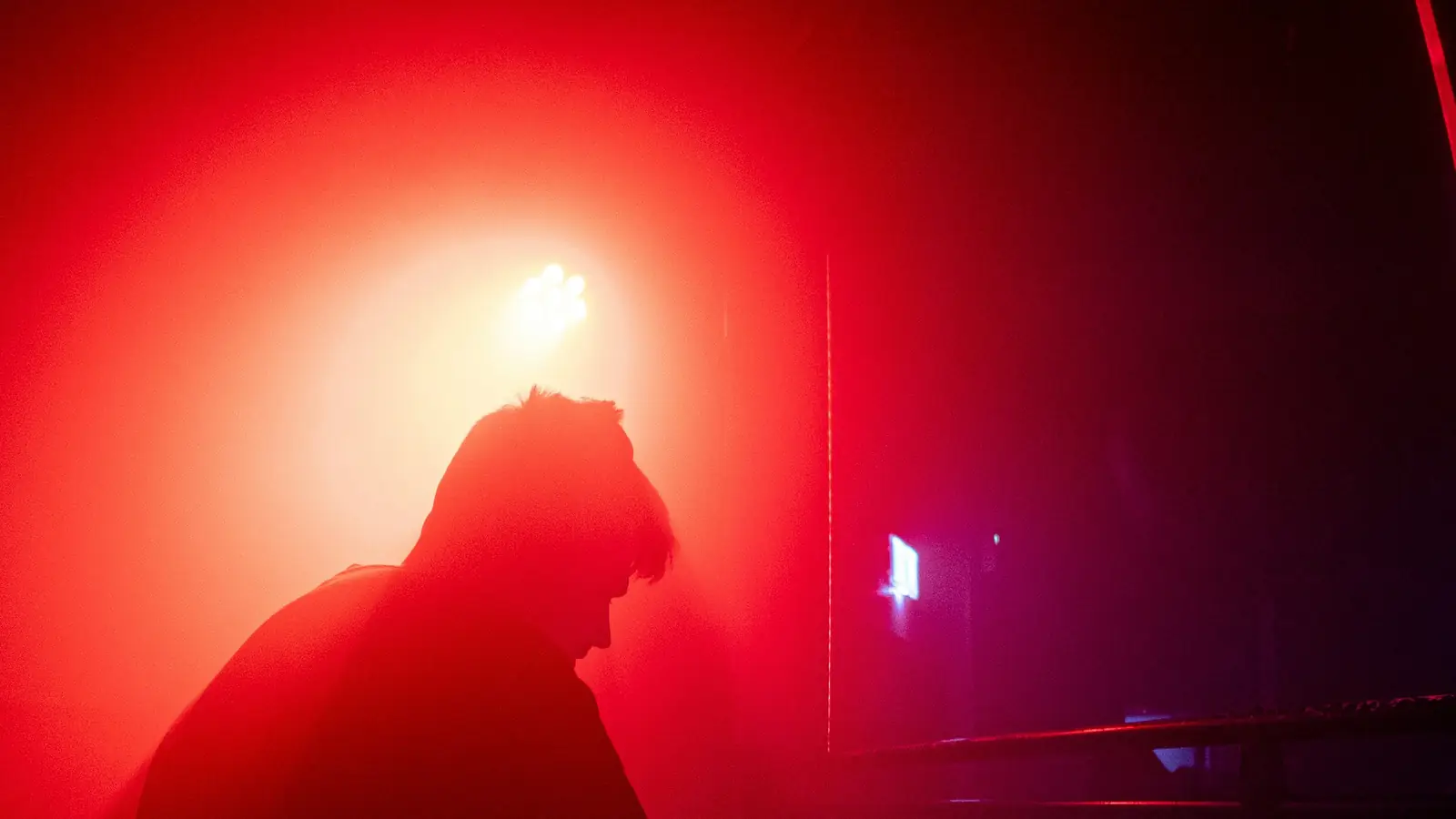 A DJ in silhouette against a wash of red light, head bowed over the decks, the rest of the room dark and out of frame