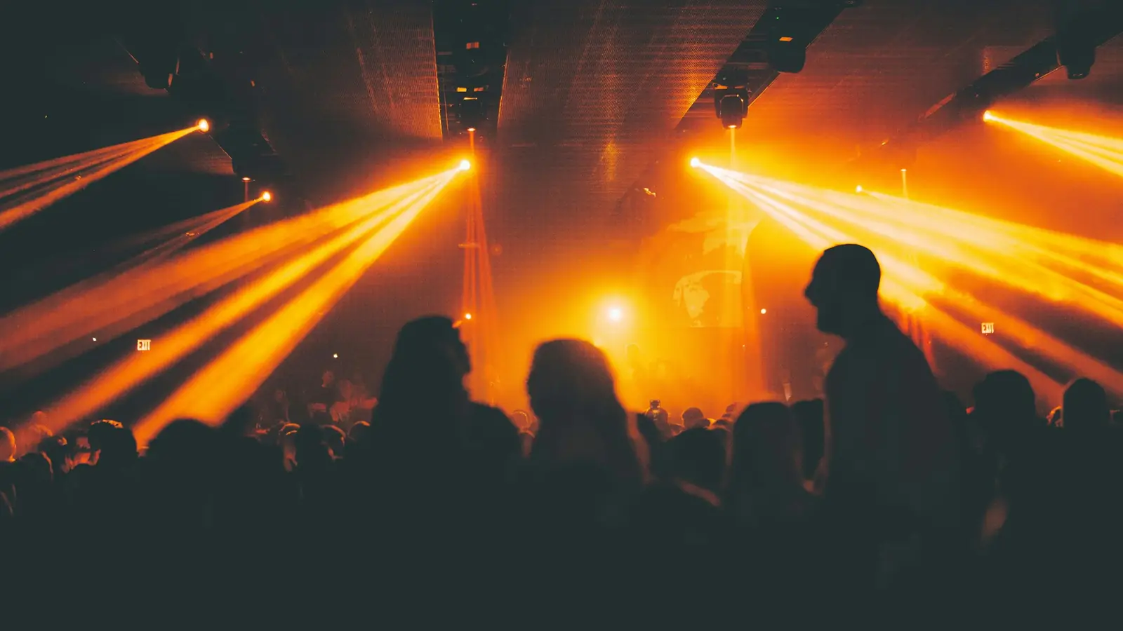 A crowd in silhouette under fanning amber beams, mid-set, the room already committed rather than deciding