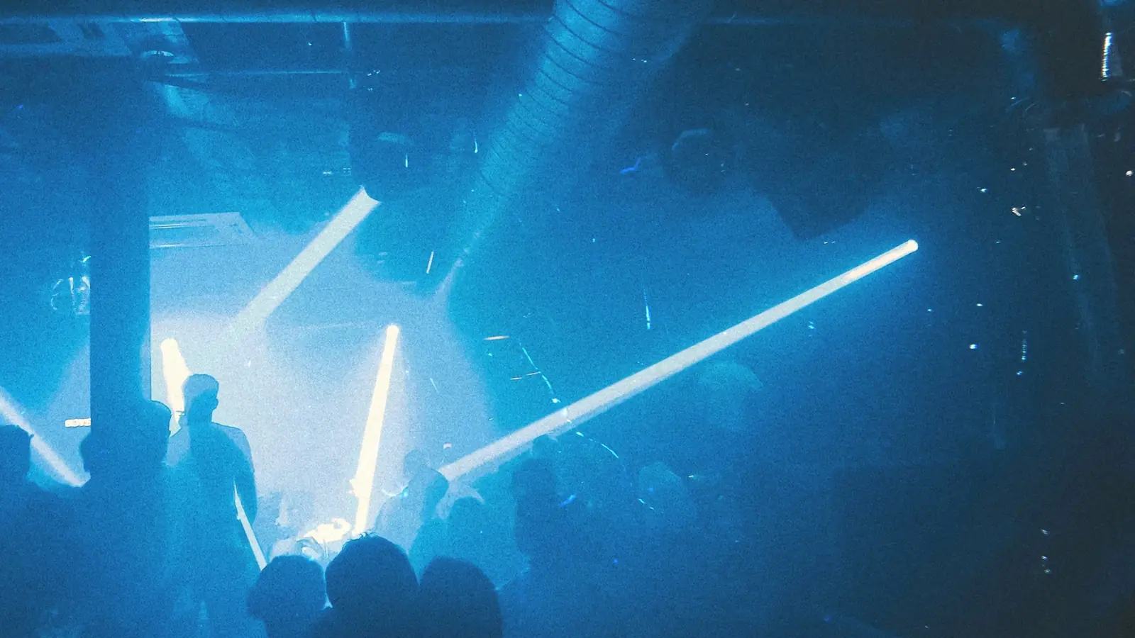 A packed, hazy room lit in deep blue, beams of light crossing over the silhouettes of a crowd locked into the same direction