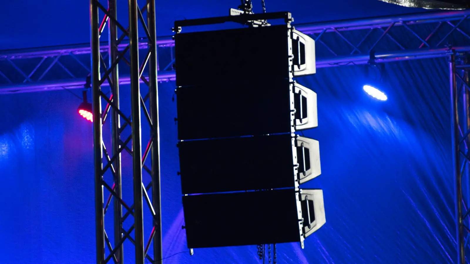 A stacked line-array speaker cluster hangs from a rigging truss, lit in deep blue with one red beam crossing it, against a dark night sky