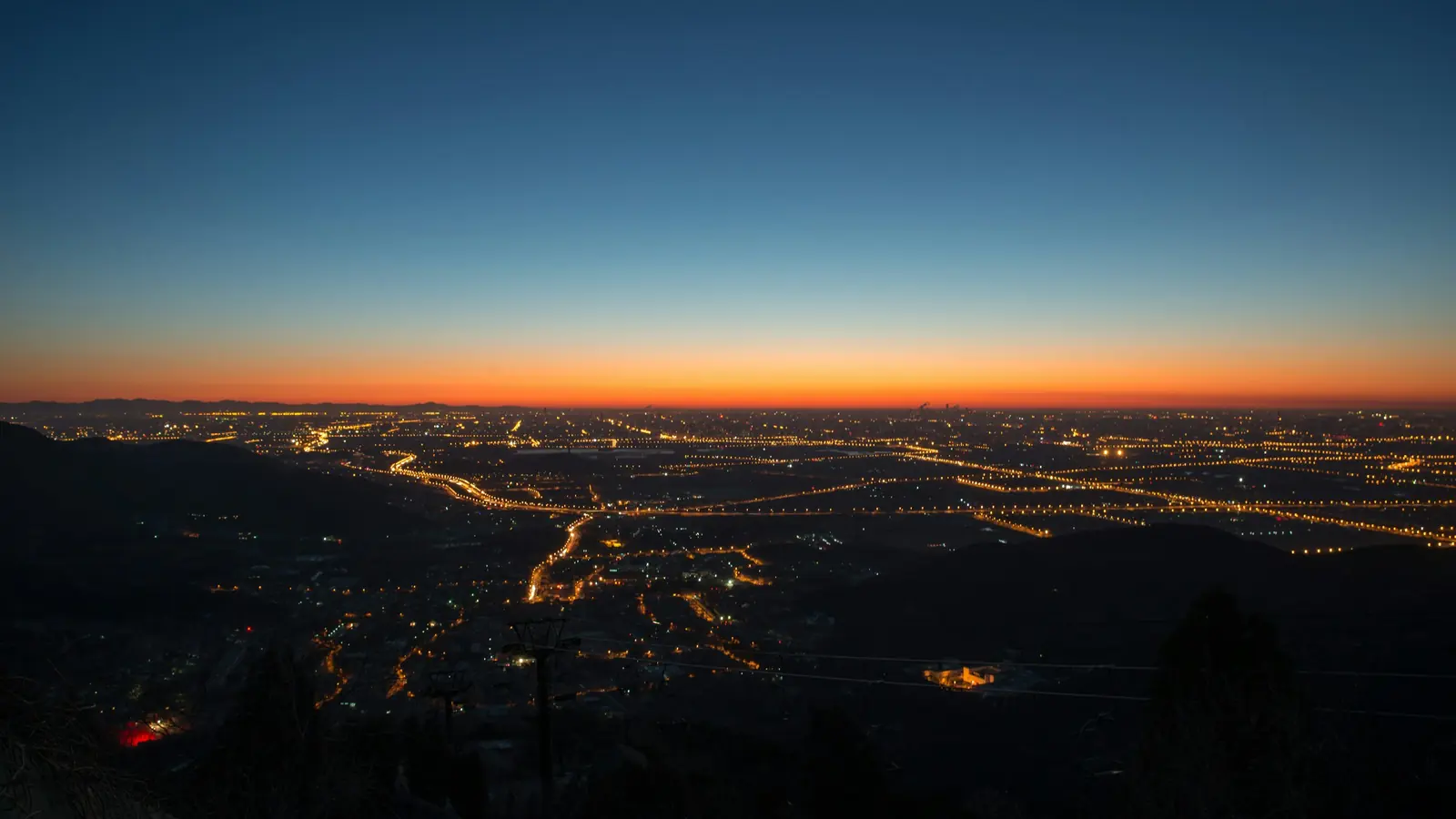 A city seen from high ground at twilight, street lights burning in long orange lines beneath a band of light on the horizon and deep blue sky above
